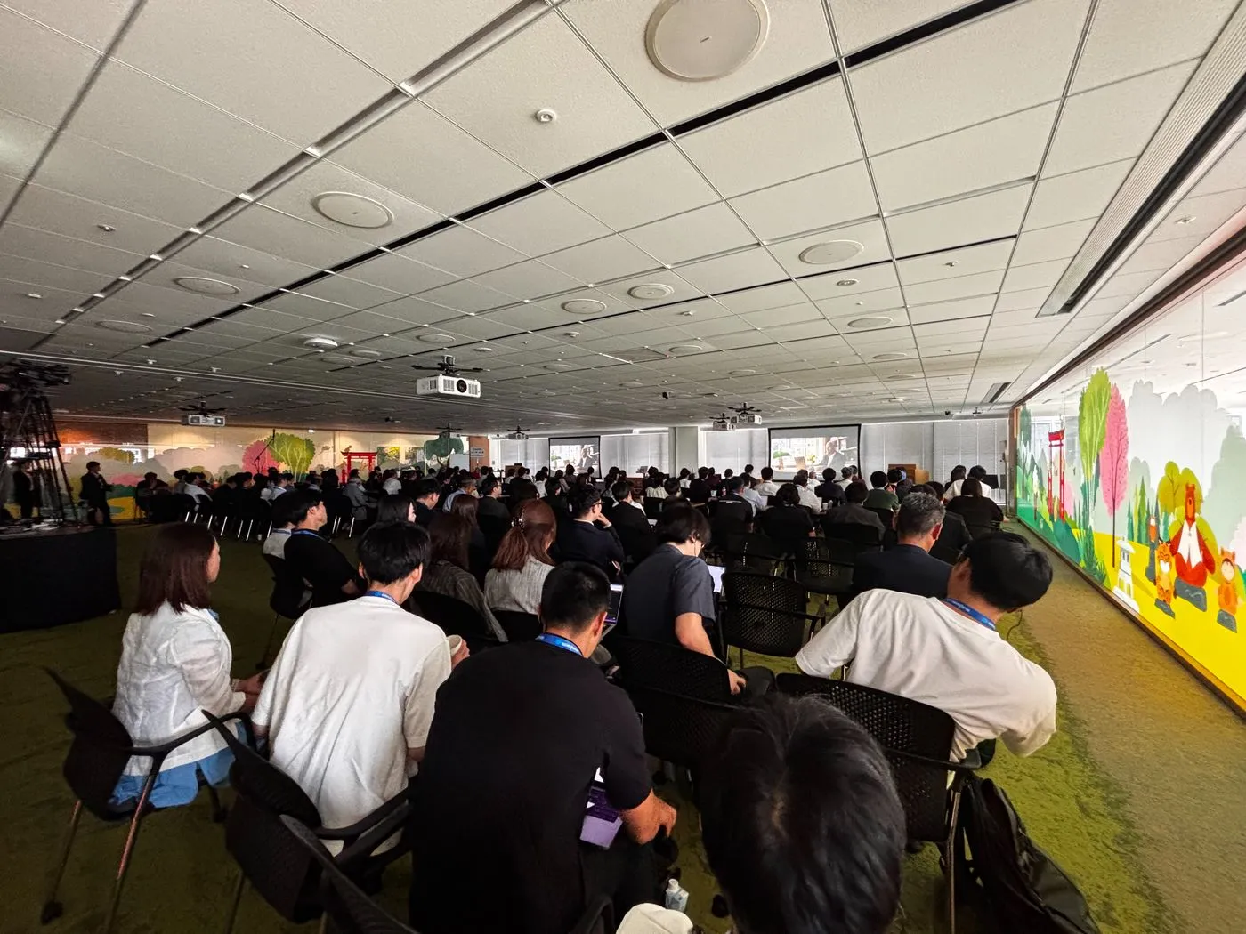 Salesforce Tower Tokyo のセミナールームで開催された Marketing & Commerce Innovation Day の会場全景。緑のカーペットとイラストの壁面に囲まれた会場に参加者が着席し、正面のスクリーンで基調講演が進んでいる